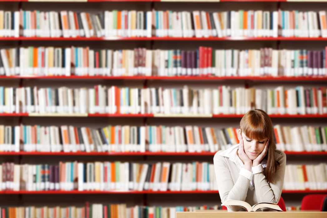 Bild vergrößern: Das Bild zeigt eine junge Frau, die in der Bibliothek ein Buch liest.