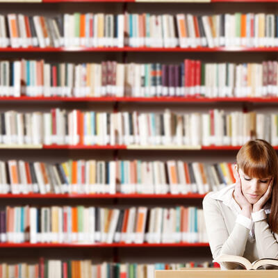Das Bild zeigt eine junge Frau, die in der Bibliothek ein Buch liest.