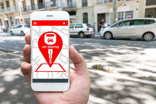 Bild vergrößern: Car sharing service or rental concept. Sharing economy and collaborative consumption. Customer hand holding smart phone with icons application screen and blur car background.