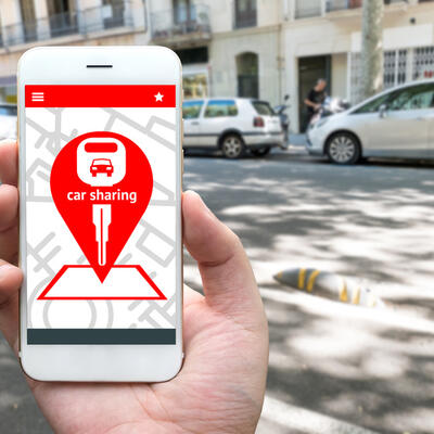 Bild vergrößern: Car sharing service or rental concept. Sharing economy and collaborative consumption. Customer hand holding smart phone with icons application screen and blur car background.