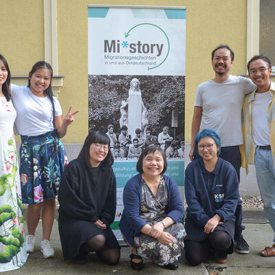 Asiatische Frauen und Männer vor einem Roll-Up "Migrationsgeschichten"